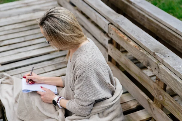 En blond kvinna sitter, täckt med en filt, ritar en skiss eller skriver en bok. — Stockfoto