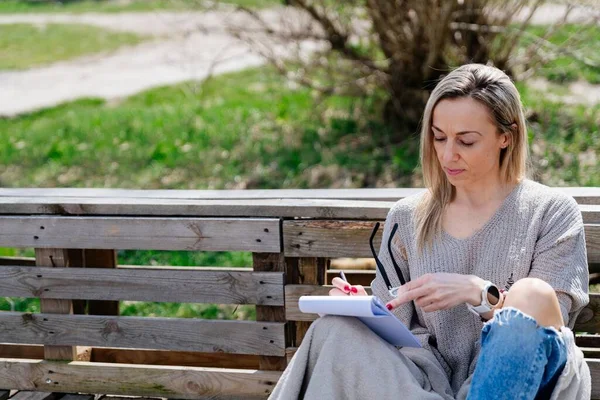 Voyageuse, écrivaine assise sur un banc à palettes réfléchissant et écrivant ou dessinant quelque chose dans un carnet. — Photo