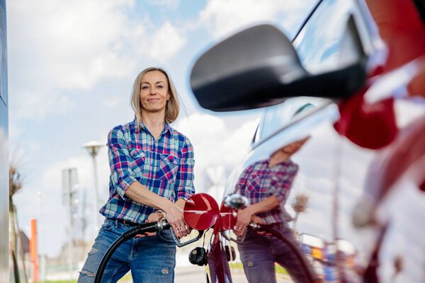 Blonde woman is filling her red car with gasoline at a gas station.