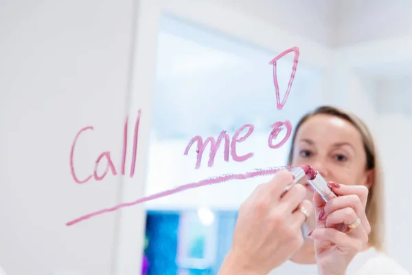 Donna bionda scrive le parole chiamami sullo specchio con rossetto rosso. — Foto Stock