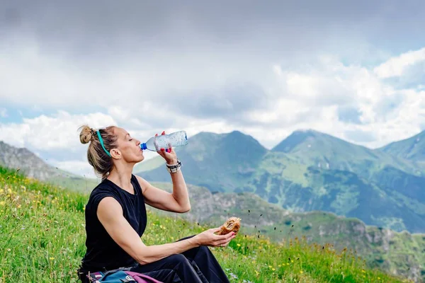 Femme randonneuse assise, buvant de l'eau et mangeant un sandwich, reposant sur l'herbe dans les montagnes. — Photo