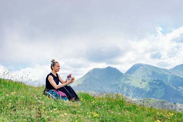 A blonde tourist sits on the grass, eats and drinks water, rests in the beautiful tatra mountain. — Stock Photo, Image