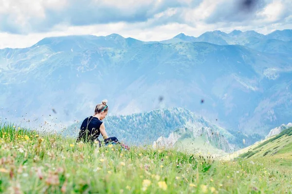 Vue arrière d'une voyageuse blonde reposant sur l'herbe lors d'un trek en montagne. — Photo