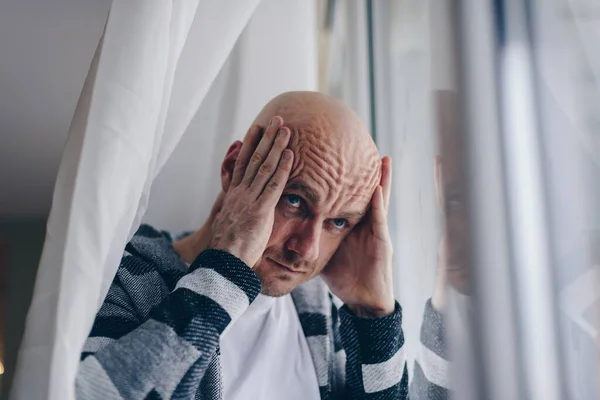 Un hombre triste sostiene su cabeza con las manos, mira por la ventana. — Foto de Stock