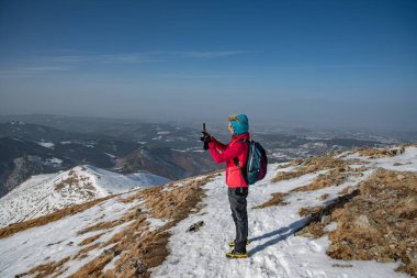 A tourist in a red jacket and backpack takes a photo with a phone on a beautiful mountain trail.