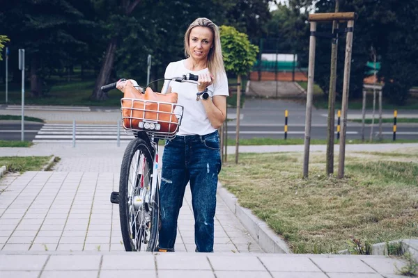 A worried woman in a white T-shirt climbs the stairs with a city bike. — Stock Fotó