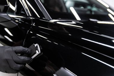 Parlak siyah araba boyasına elle uygulanan seramik kaplamanın yakın çekimi, derin yansımaları vurgulamak, hassas teknik, boya koruması ve özel araba temizleme servisi.