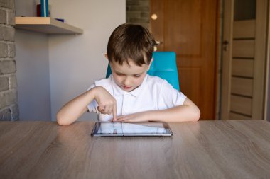 Masada otururken tablet kullanan 6 yaşındaki çocuk. Beyaz polo gömlek giymiş