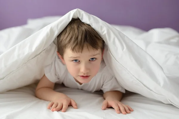 7-jähriger Junge versteckt sich im Bett unter einer weißen Decke oder Decke — Stockfoto