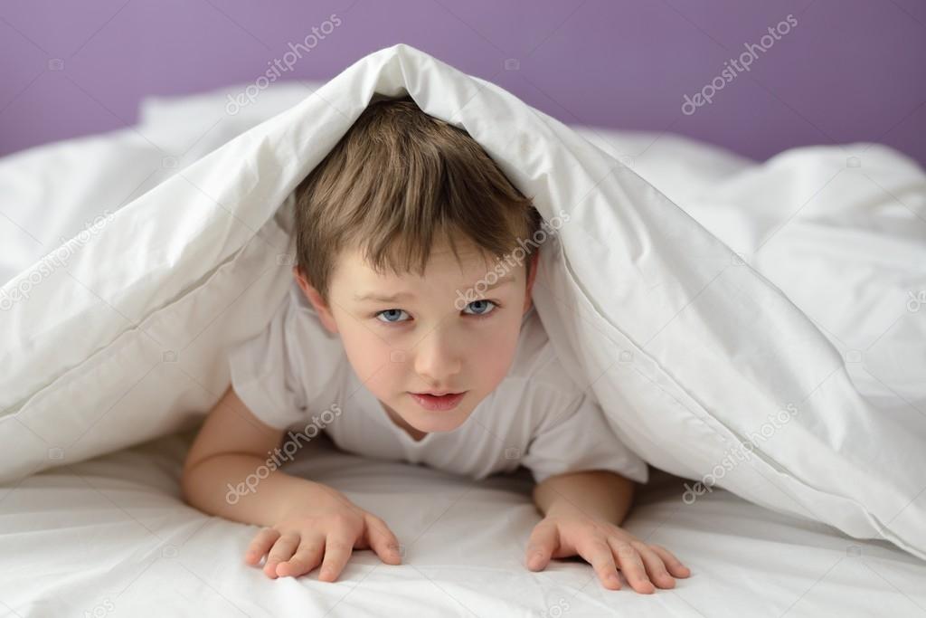 7 years old boy hiding in bed under a white blanket or coverlet Stock