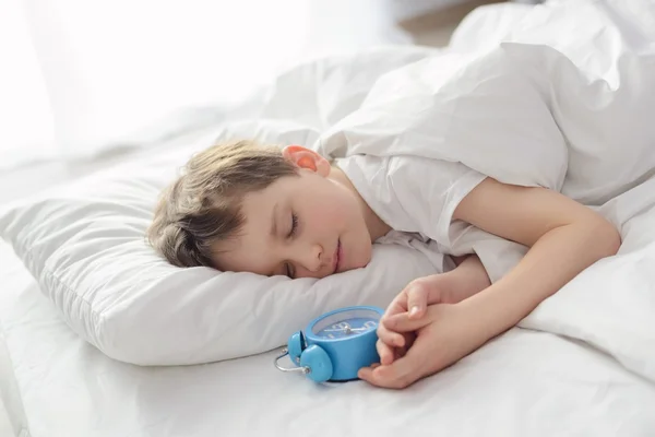 Niño durmiendo en cama blanca con despertador cerca de su cabeza —  Fotos de Stock