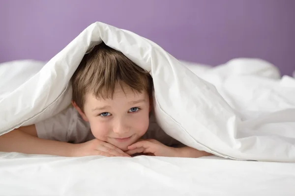 Menino feliz escondido na cama sob um cobertor branco ou coverlet — Fotografia de Stock