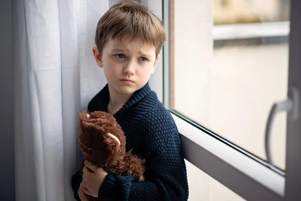 O rapaz está a abraçar o ursinho. Ao pé da janela . — Fotografia de Stock
