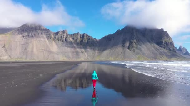 大西洋沿岸をジョギング中のアクティブな女性の空中ビュー 海の波黒い砂浜を洗う ホフン ヴェストラホーン山脈 アイスランド — ストック動画