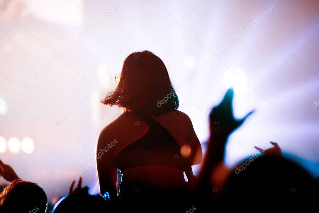 Rear view of crowd with arms outstretched at concert. Bright stage lights in the background