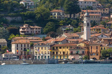 Menaggio, İtalya - 21 Ağustos 2021: Como Gölü 'nden geleneksel binalar ve liman üzerinde yükselen kilise ile birlikte Menaggio köyünün görüntüsü.