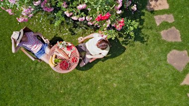 Romantik bir buluşmada, güzel gül bahçesinde yemek ve içeceklerin keyfini çıkaran genç bir çift. Parkta birlikte yiyip içen erkek ve kadının yukarıdan görünüşü.