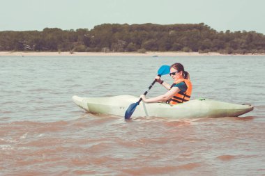 Deniz, göl, geniş nehir. Can yeleği giymiş bir kız kürek çekiyor. Uzak kıyılar, ağaçlar. Sıcak yaz günü, güneşli aydınlatma. Tonlama.