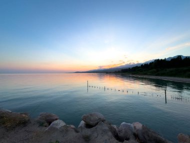 Kırgızistan 'daki Issyk Kul Gölü' nde gün batımı. Güneş ışınları bulutları delip geçiyor. Sakin sulara yansıyor ve arka planda dağlar var. Issyk Kul 'da gün batımında güneşin yansıması ve Tien Shan dağlarında.