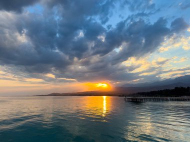 Kırgızistan 'daki Issyk Kul Gölü' nde gün batımı. Parlak güneş bulutları delip geçiyor ve turkuaz suyu yansıtıyor. Gün batımında Issyk Kul 'un turkuaz suyu altın yansıma ve dramatik bulutlarla.