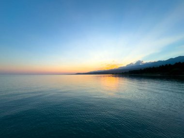 Kırgızistan 'daki Issyk Kul Gölü' nde gün batımı. Güneş ışınları bulutları delip geçiyor. Sakin sulara yansıyor ve arka planda dağlar var. Issyk Kul 'da gün batımında güneşin yansıması ve Tien Shan dağlarında.