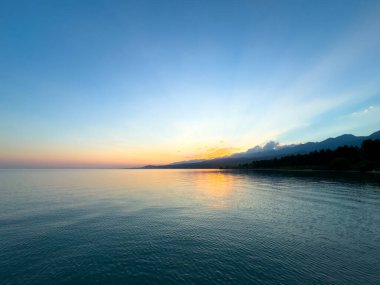 Kırgızistan 'daki Issyk Kul Gölü' nde gün batımı. Güneş ışınları bulutları delip geçiyor. Sakin sulara yansıyor ve arka planda dağlar var. Issyk Kul 'da gün batımında güneşin yansıması ve Tien Shan dağlarında.