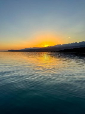Kırgızistan 'daki Issyk Kul Gölü üzerinde gün batımı. Güneş dağların arkasında batar, suyun üzerinde altın bir yansıma bırakır. Kırgızistan 'daki Issyk Kul Gölü üzerinde gün batımında suyun üzerinde altın yansımasıyla.