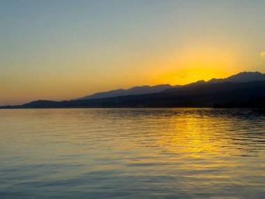 Kırgızistan 'daki Issyk Kul Gölü üzerinde gün batımı. Güneş dağların arkasında batar, suyun üzerinde altın bir yansıma bırakır. Kırgızistan 'daki Issyk Kul Gölü üzerinde gün batımında suyun üzerinde altın yansımasıyla.