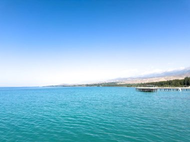 Kırgızistan 'da güneşli bir günde Issyk Kul Gölü. Turkuaz su, uzun iskele ve ufukta Tien Shan dağları. Issyk Kul 'un turkuaz suyu iskele ve Tien Shan dağları açık bir günde.