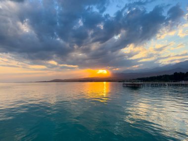 Kırgızistan 'daki Issyk Kul Gölü' nde gün batımı. Parlak güneş bulutları delip geçiyor ve turkuaz suyu yansıtıyor. Gün batımında Issyk Kul 'un turkuaz suyu altın yansıma ve dramatik bulutlarla.