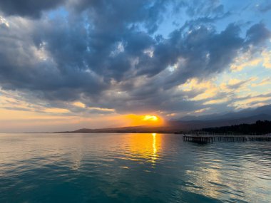 Kırgızistan 'daki Issyk Kul Gölü' nde gün batımı. Parlak güneş bulutları delip geçiyor ve turkuaz suyu yansıtıyor. Gün batımında Issyk Kul 'un turkuaz suyu altın yansıma ve dramatik bulutlarla.