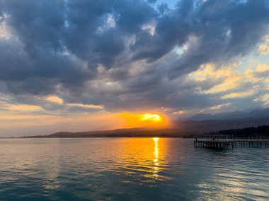 Kırgızistan 'daki Issyk Kul Gölü' nde gün batımı. Parlak güneş bulutları delip geçiyor ve turkuaz suyu yansıtıyor. Gün batımında Issyk Kul 'un turkuaz suyu altın yansıma ve dramatik bulutlarla.