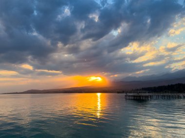 Kırgızistan 'daki Issyk Kul Gölü' nde gün batımı. Parlak güneş bulutları delip geçiyor ve turkuaz suyu yansıtıyor. Gün batımında Issyk Kul 'un turkuaz suyu altın yansıma ve dramatik bulutlarla.