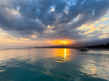 Kırgızistan 'daki Issyk Kul Gölü' nde gün batımı. Parlak güneş bulutları delip geçiyor ve turkuaz suyu yansıtıyor. Gün batımında Issyk Kul 'un turkuaz suyu altın yansıma ve dramatik bulutlarla.