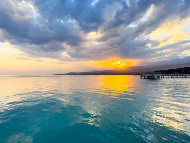 Kırgızistan 'daki Issyk Kul Gölü' nde gün batımı. Parlak güneş bulutları delip geçiyor ve turkuaz suyu yansıtıyor. Gün batımında Issyk Kul 'un turkuaz suyu altın yansıma ve dramatik bulutlarla.