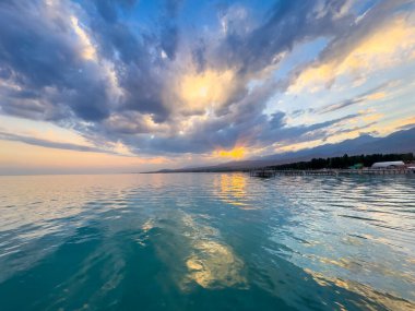 Kırgızistan 'daki Issyk Kul Gölü' nde gün batımı. Parlak güneş bulutları delip geçiyor ve turkuaz suyu yansıtıyor. Gün batımında Issyk Kul 'un turkuaz suyu altın yansıma ve dramatik bulutlarla.