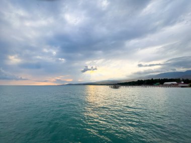 Kırgızistan 'daki Issyk Kul Gölü. Sakin turkuaz su, bulutları yansıtıyor. Plaj ve rıhtımın üzerinde yumuşak akşam ışığı var. Issyk Kul 'un turkuaz suyu ve akşam ışığında kıyı şeridinde çarpıcı bir gökyüzü.