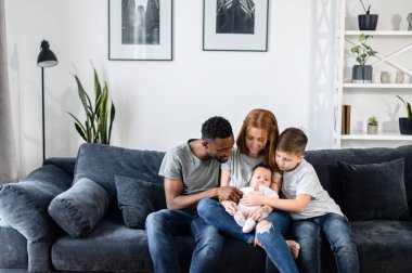 A multiracial family sits on the couch at home clipart