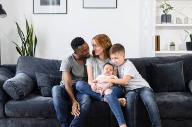 A multiracial family sits on the couch at home clipart