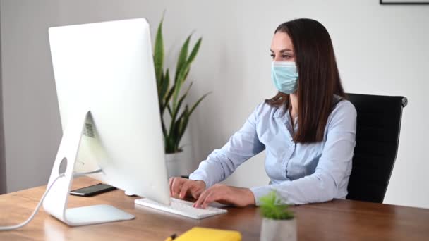 Eine Frau in Maske mit PC im Büro — Stockvideo