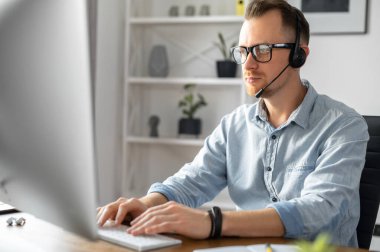 Ofiste kulaklık takan, modern bilgisayar kullanan, internette konuşan ve masada oturan genç bir erkeğin yan görüntüsü. Çağrı merkezi, yardım hattı, destek servisi