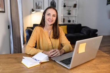 Çevrimiçi konuşmak için kulaklık kullanan neşeli genç bir kadın merkez ofisteki masada oturuyor, hoş bayan operatör kameraya bakıyor. Uzak çalışma konsepti