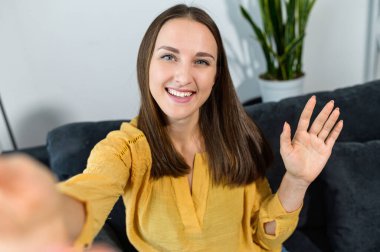 Neşeli genç bir kadın akıllı bir telefon tutuyor, webcam 'e el sallıyor, gülümsüyor ve selamlıyor. Video görüşmesi, video konferansı, online toplantı. Çekici bir kızla görüntülü sohbet