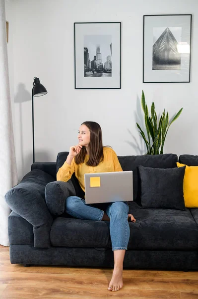 Dreaming young woman sits on a sofa with a laptop. Smiling girl in casual wear looks away lost in pleasant thoughts, relaxing on comfortable couch in a cozy living room, takes a break