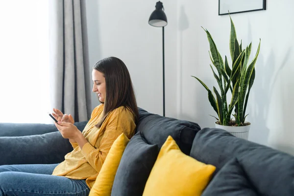 Вид сбоку спокойная женщина расслабляется на удобном диване, держа в руках смартфон, общаясь в социальных сетях, смс и прокручивая ленту новостей. Девушка проводит досуг с мобильным телефоном — стоковое фото