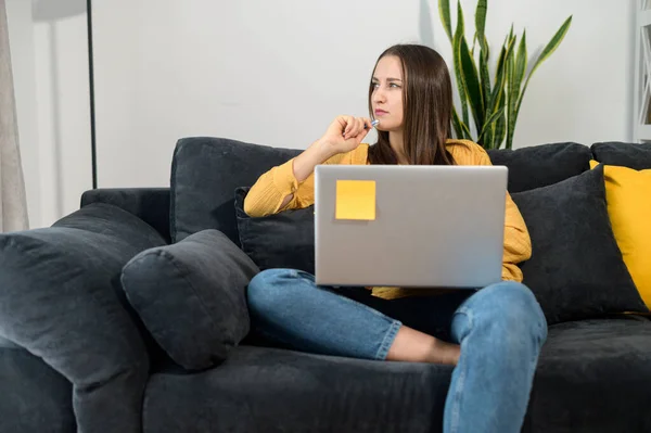 Удумлива жінка-підприємець дивиться вбік, сидячи на зручному дивані з ноутбуком. Фрілансер жінка в повсякденному вбранні працює в Інтернеті віддалено — стокове фото
