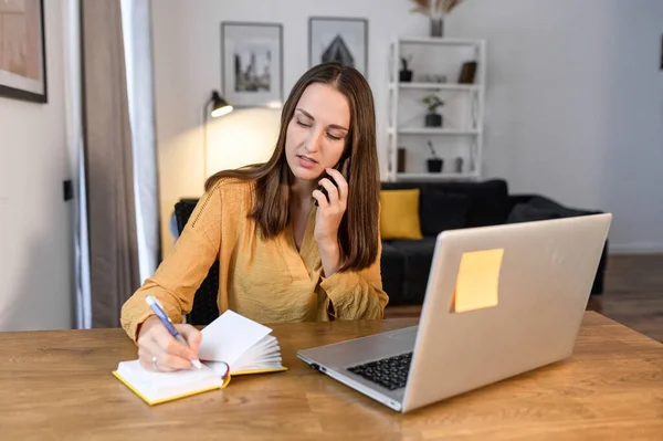 Молодая женщина-фрилансер разговаривает по телефону и использует ноутбук, работающий удаленно в домашнем офисе. Женщина в жёлтой рубашке записывает разговор по телефону. — стоковое фото