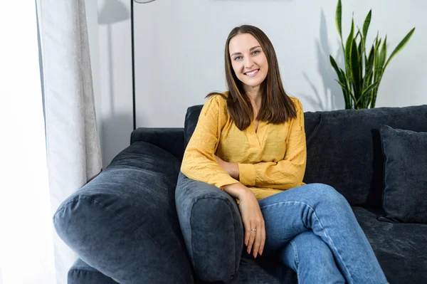 Young cheerful woman sits on a sofa at home in calm atmosphere. Smiling girl in casual wear looks into camera, relaxing on comfortable couch in a cozy living room — Stock Photo, Image