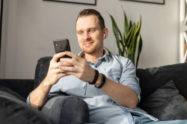 Un tipo con un teléfono inteligente se sienta en el sofá — Foto de Stock