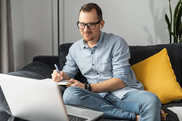 En ung man med en bärbar dator som skriver i en anteckningsbok — Stockfoto